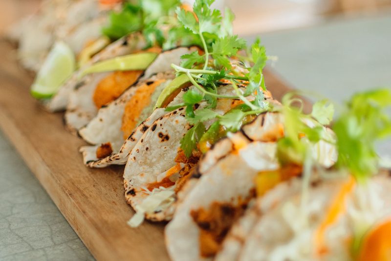 Fila de tacos recién hechos sobre tabla de madera, con cilantro, aguacate y limón